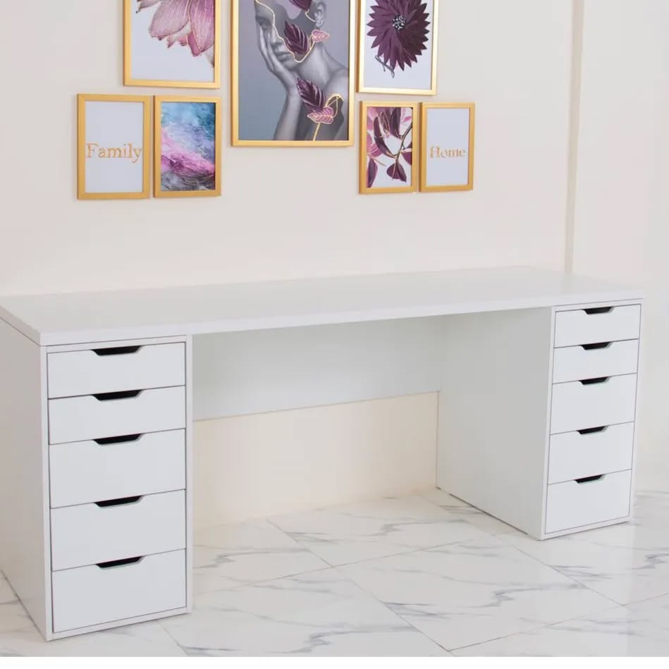 White desk with multiple drawers and framed pictures on a white wall.